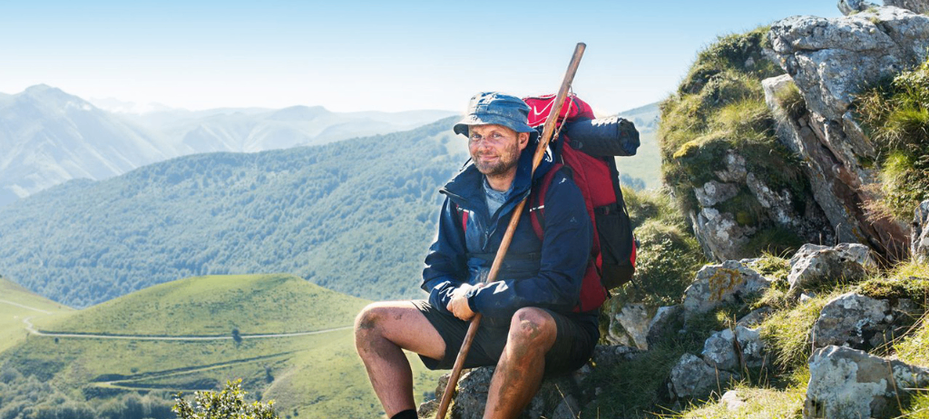 Ein Mann in blauer Jacke und Hut sitzt auf einem felsigen Berg mit Blick auf grüne Hügel und blauen Himmel. Er hält einen Stock und hat einen Rucksack auf.