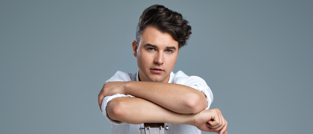 Ein Mann in einem weißen Hemd posiert mit einer Gitarre auf dem Schoß, Arme verschränkt. Er sieht ernst aus und seine Augen sind auf etwas vor ihm gerichtet.