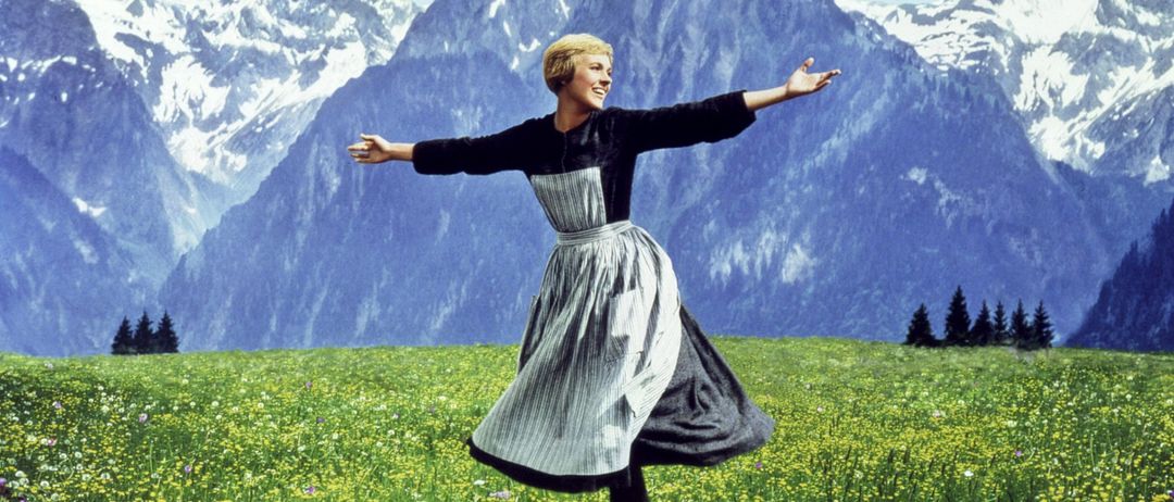 Eine Frau steht in einem Feld voller gelber Blumen mit ausgestreckten Armen vor einem Hintergrund von schneebedeckten Bergen.