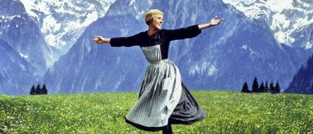 Eine Frau steht in einem Feld voller gelber Blumen mit ausgestreckten Armen vor einem Hintergrund von schneebedeckten Bergen.