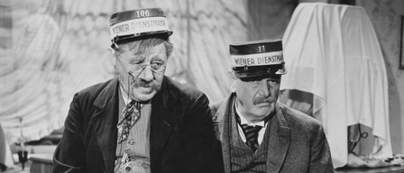 Zwei ältere Männer sitzen nebeneinander und tragen beide Uniformen. Einer ist mit 'Wiener Dienstmann 106' und der andere mit 'Wiener Dienstmann 11' beschriftet. Beide haben ernste Gesichtsausdrücke.