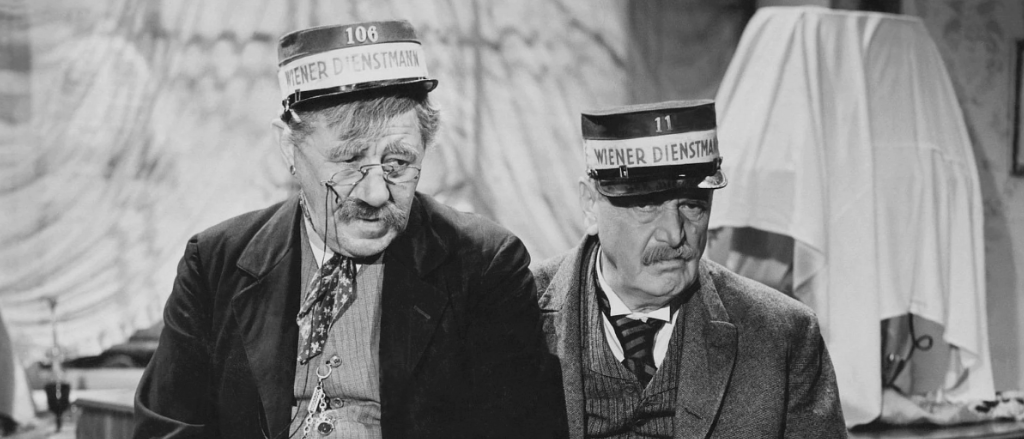 Zwei ältere Männer sitzen nebeneinander und tragen beide Uniformen. Einer ist mit 'Wiener Dienstmann 106' und der andere mit 'Wiener Dienstmann 11' beschriftet. Beide haben ernste Gesichtsausdrücke.