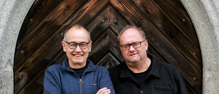 Two men are standing next to each other, smiling and posing for a photo. The man on the left wears a blue jacket and glasses. The man on the right wears a black shirt and glasses. They are in front of a wooden door.