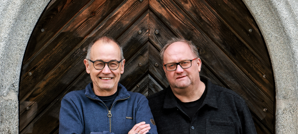 Zwei Männer stehen nebeneinander, lächeln und posieren für ein Foto. Der Mann auf der linken Seite trägt eine blaue Jacke und eine Brille. Der Mann auf der rechten Seite trägt ein schwarzes Hemd und eine Brille. Sie stehen vor einer Holztür.