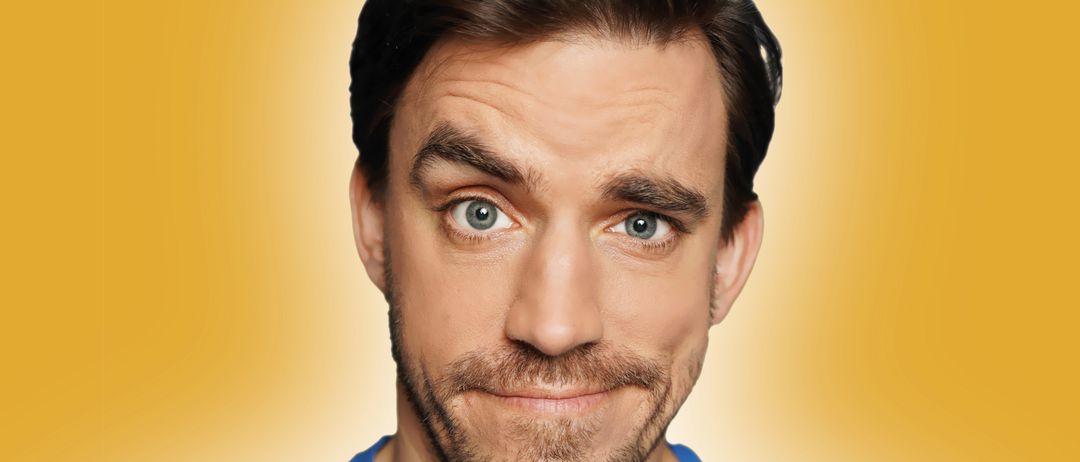 Ein Mann mit Bart und Schnurrbart, der ein blaues T-Shirt vor gelbem Hintergrund trägt.