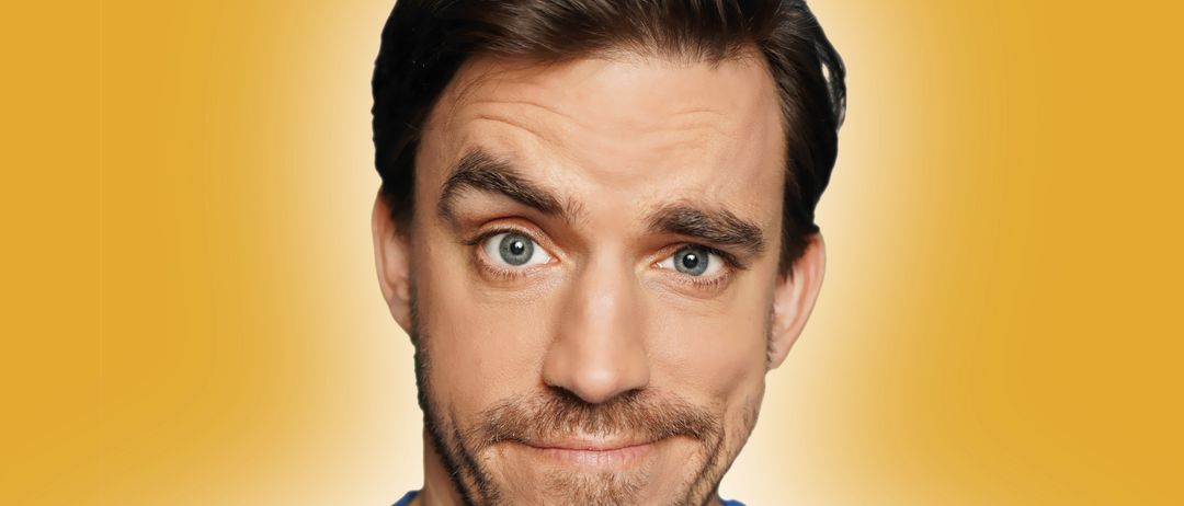 Ein Mann mit Bart und Schnurrbart in einem blauen T-Shirt lächelt in die Kamera vor einem hellen gelben Hintergrund.