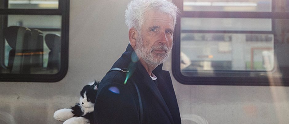 Ein älterer Mann mit weißen Haaren und Bart steht vor einem Zug und trägt ein Kuscheltier auf der Schulter.