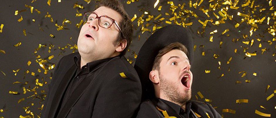 Zwei Männer in schwarzen Anzügen und Hüten, posieren für ein Foto mit goldenem Konfetti, das im Hintergrund fällt.