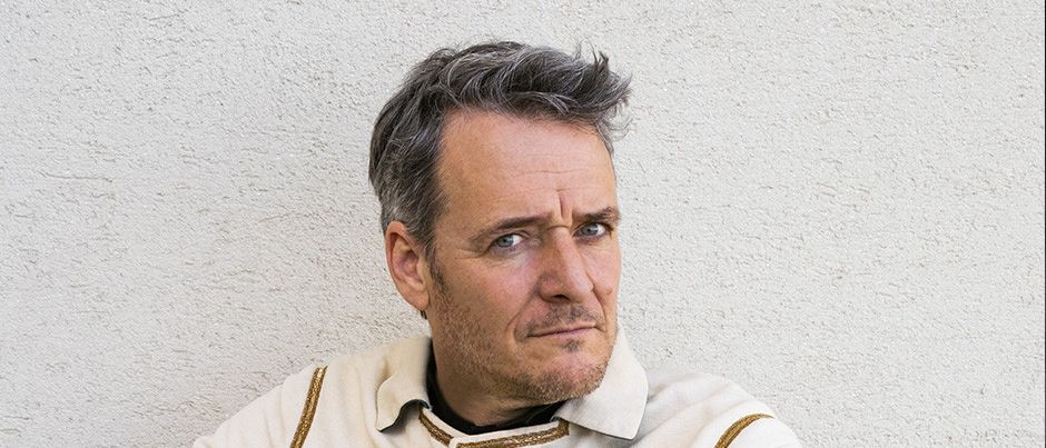 Ein Mann mit grauem Haar und Bart trägt eine weiße Jacke mit goldenen Verzierungen und Knöpfen. Er sitzt auf einem Holzstuhl und berührt seine Brust mit der linken Hand.