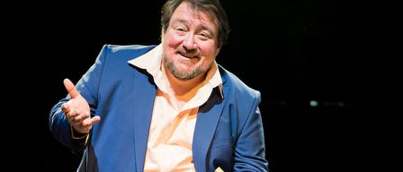Ein Mann in einem blauen Anzug und Hosen steht auf der Bühne, hält ein gelbes Buch und lächelt während einer Aufführung.