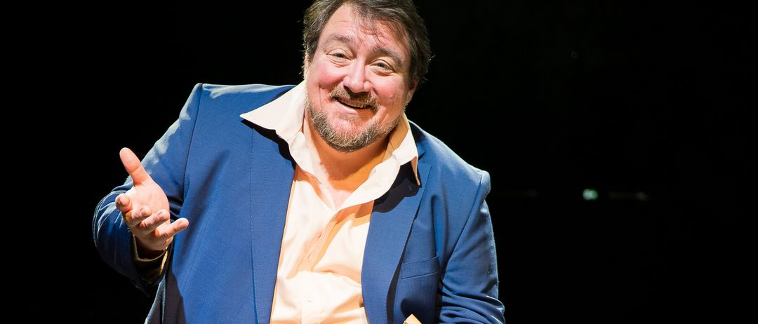 Ein Mann in einem blauen Anzug und Hosen steht auf der Bühne, hält ein gelbes Buch und lächelt während einer Aufführung.
