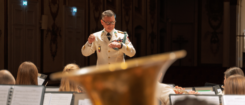 Ein Mann in Uniform dirigiert ein Orchester, mit Musikern, die im Vordergrund Instrumente spielen.