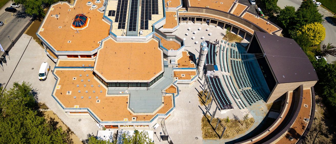 Luftaufnahme eines modernen architektonischen Gebäudes mit einem großen sechseckigen Dach, das Solarmodule aufweist. Die Struktur ist von einem halbkreisförmigen Sitzbereich und Grünflächen umgeben.