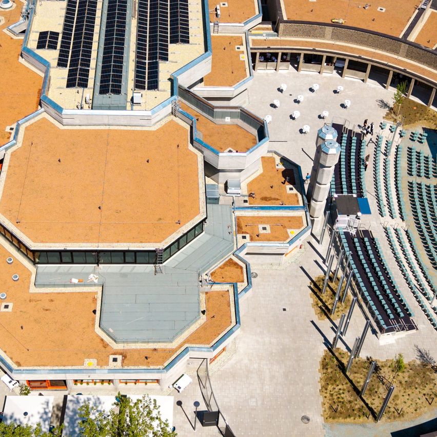 Luftaufnahme eines modernen architektonischen Gebäudes mit einem großen sechseckigen Dach, das Solarmodule aufweist. Die Struktur ist von einem halbkreisförmigen Sitzbereich und Grünflächen umgeben.