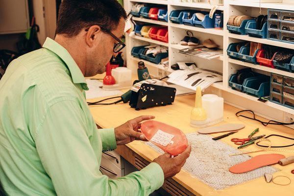 Ein Mann mit Brille und grünem Hemd arbeitet an einem rosa Objekt, möglicherweise einer Schuhsohle, auf einem Schreibtisch mit verschiedenen Werkzeugen und Materialien.