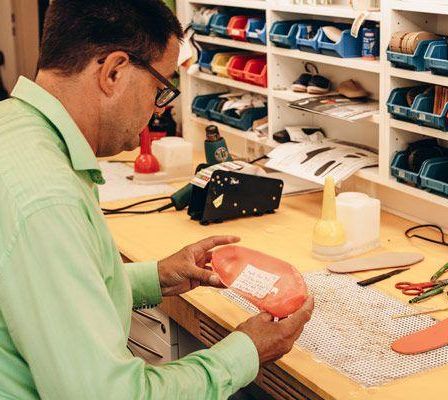 Ein Mann mit Brille und grünem Hemd arbeitet an einem rosa Objekt, möglicherweise einer Schuhsohle, auf einem Schreibtisch mit verschiedenen Werkzeugen und Materialien.