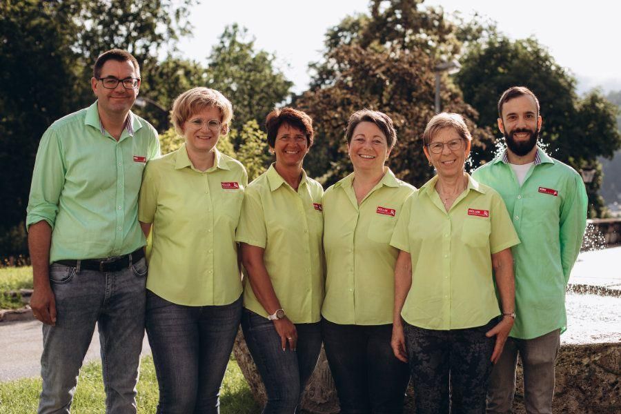 Sechs Menschen in passenden hellgrünen Hemden stehen in einer Reihe vor einem Hintergrund aus Bäumen und Gras. Drei Frauen und drei Männer, alle lächeln.