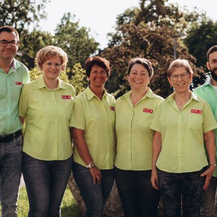 Sechs Menschen in passenden hellgrünen Hemden stehen in einer Reihe vor einem Hintergrund aus Bäumen und Gras. Drei Frauen und drei Männer, alle lächeln.