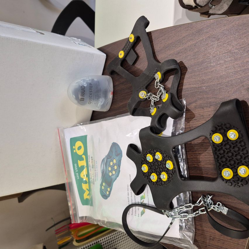 A wooden table displays a pair of black and yellow crampons, a small plastic bottle, a white box, and a white package.