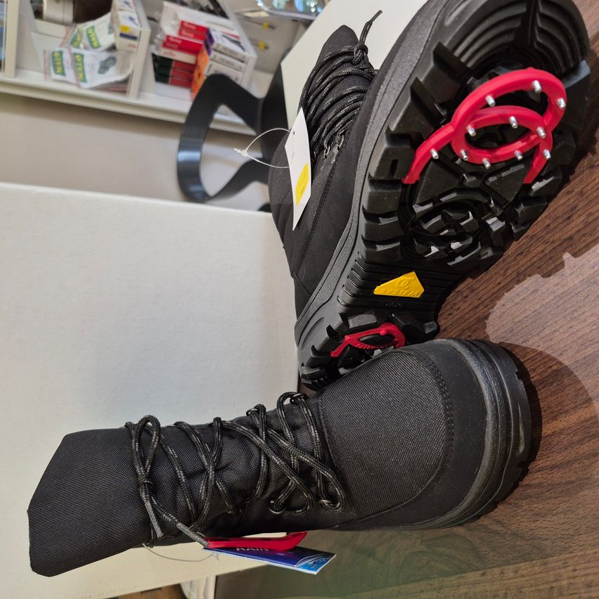 A pair of black winter boots with red and yellow accents on a wooden surface, with a tag hanging from the laces. Behind them is a shelf with various items.