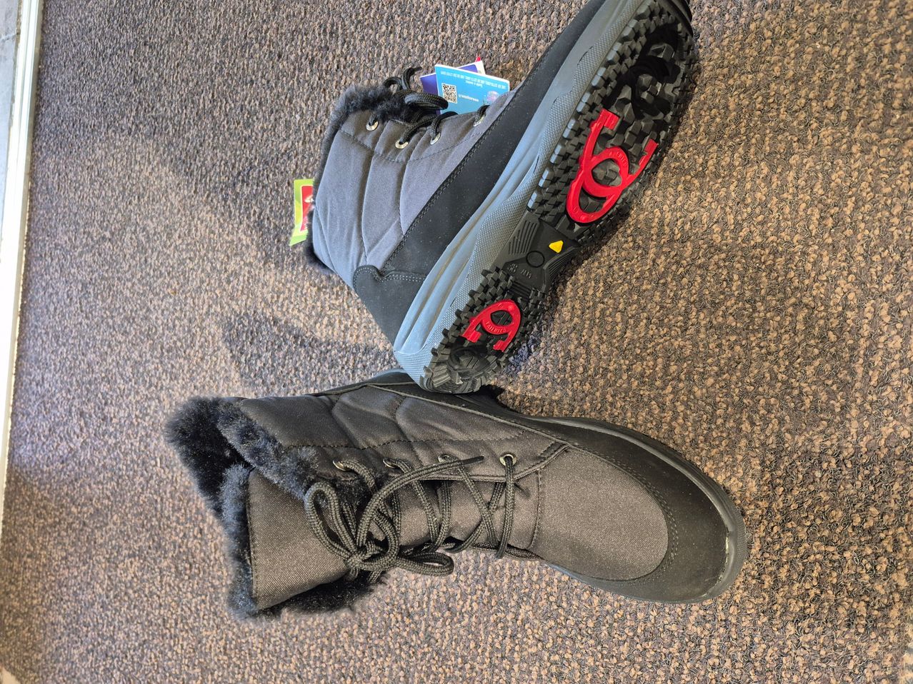 A pair of dark gray and black boots with fur lining, laces, and red number '6' on the soles, placed on a brown carpet.
