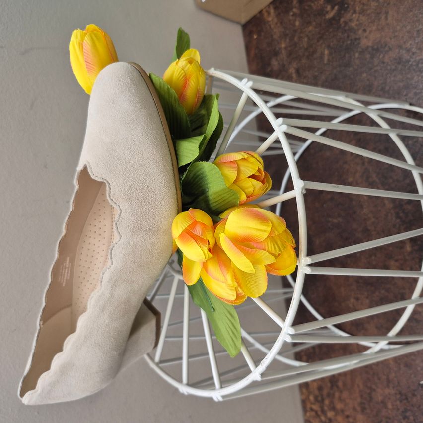 Ein beigefarbener Schuh mit gezackten Kanten steht auf einem weißen Drahtständer mit gelben Tulpen. Die Tulpen sind dekorativ im Schuh angeordnet.