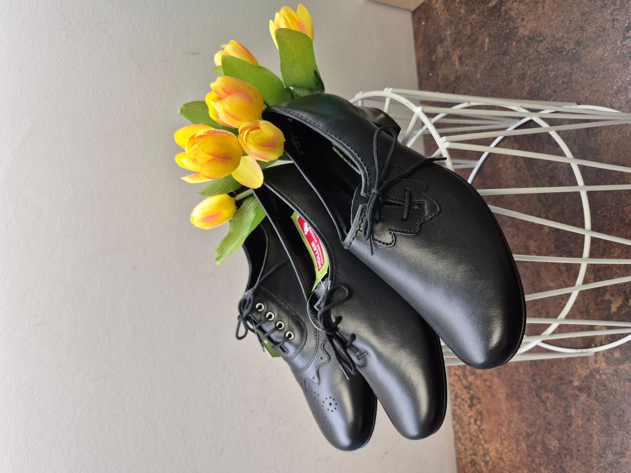 Ein Paar schwarze Lederschuhe mit gelben Tulpen auf einem weißen Korb. Die Schuhe haben einen Aufkleber an der Seite.