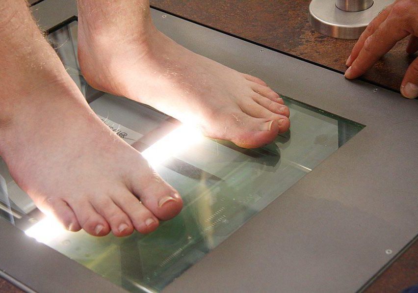 A person's feet are on a digital scale, with a hand touching the side of the scale. The feet are illuminated by a light, and the scale is reflecting the light.