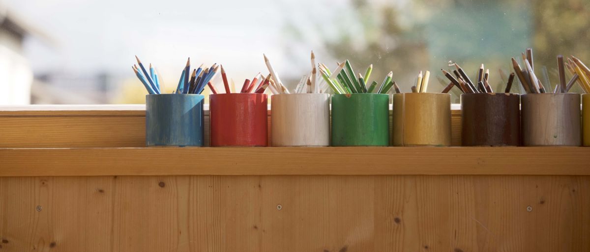 Fünf farbige Bleistifte in kleinen Behältern sind auf einem hölzernen Fensterbrett aufgereiht. Das Fenster zeigt eine verschwommene Außenansicht.