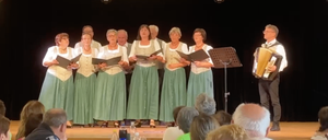 Eine Gruppe von Menschen in grünen Kleidern singt auf der Bühne mit einem Akkordeonspieler. Das Publikum sitzt vor ihnen, und es gibt Flaschen und Weingläser auf dem Tisch.