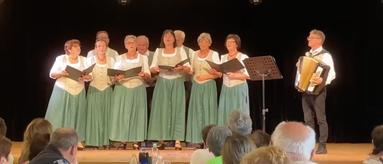 Eine Gruppe von Menschen in grünen Kleidern singt auf der Bühne mit einem Akkordeonspieler. Das Publikum sitzt vor ihnen, und es gibt Flaschen und Weingläser auf dem Tisch.