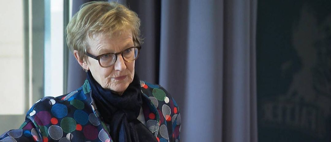 Eine ältere Frau mit kurzen blonden Haaren steht vor einem Vorhang. Sie trägt eine Brille und eine farbenfrohe Jacke. Sie hat einen ernsten Gesichtsausdruck.