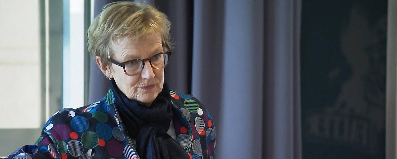 Eine ältere Frau mit kurzen blonden Haaren steht vor einem Vorhang. Sie trägt eine Brille und eine farbenfrohe Jacke. Sie hat einen ernsten Gesichtsausdruck.