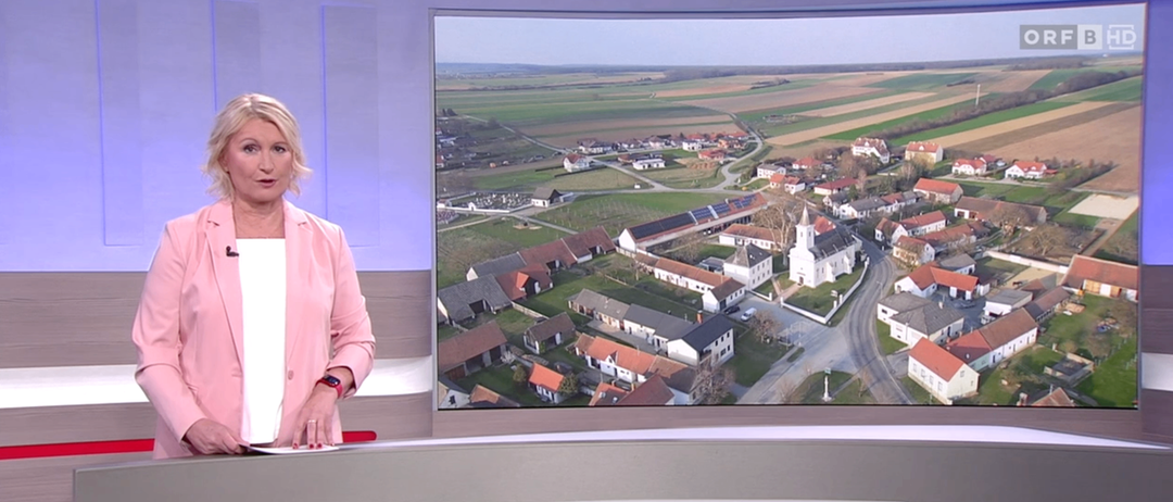 Ein Nachrichtensprecher steht mit einem Papier und zeigt eine Luftaufnahme eines Dorfes mit Häusern, grünen Feldern, einer Kirche und einer Straße.