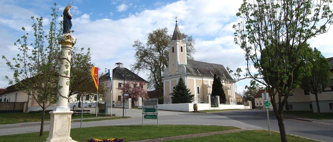 Eine Kirche mit einem Kirchturm und einem Kreuz steht neben einem Gebäude mit Fahnen und Bäumen. Eine Straße führt zur Kirche, an der ein Wegweiser auf Weinarchiv zeigt.