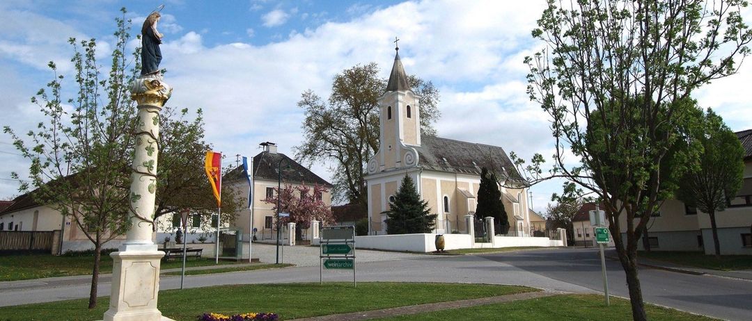 Eine alte Kirche mit einem Turm und einem Fahnenmast steht in der Nähe einer Straße. Ein Schild davor zeigt Weinarchiv. Es gibt Bäume, einen Zaun und ein Haus in der Nähe. Der Himmel ist bewölkt.