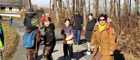 Eine Gruppe von Menschen, einschließlich Erwachsener und Kinder, geht auf einem von Bäumen gesäumten Pfad. Einige von ihnen tragen Jacken und haben Rucksäcke dabei. Eine Frau trägt eine Sonnenbrille.