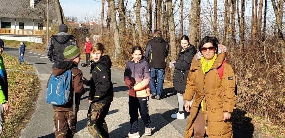 Eine Gruppe von Menschen, einschließlich Erwachsener und Kinder, geht auf einem von Bäumen gesäumten Pfad. Einige von ihnen tragen Jacken und haben Rucksäcke dabei. Eine Frau trägt eine Sonnenbrille.