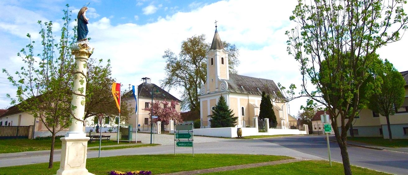 Eine Kirche mit Turm steht neben einem Gebäude mit einer roten Fahne. Straßenschilder zeigen die Richtung nach Weinarchiv. Im Vordergrund befinden sich Bäume und Blumen.