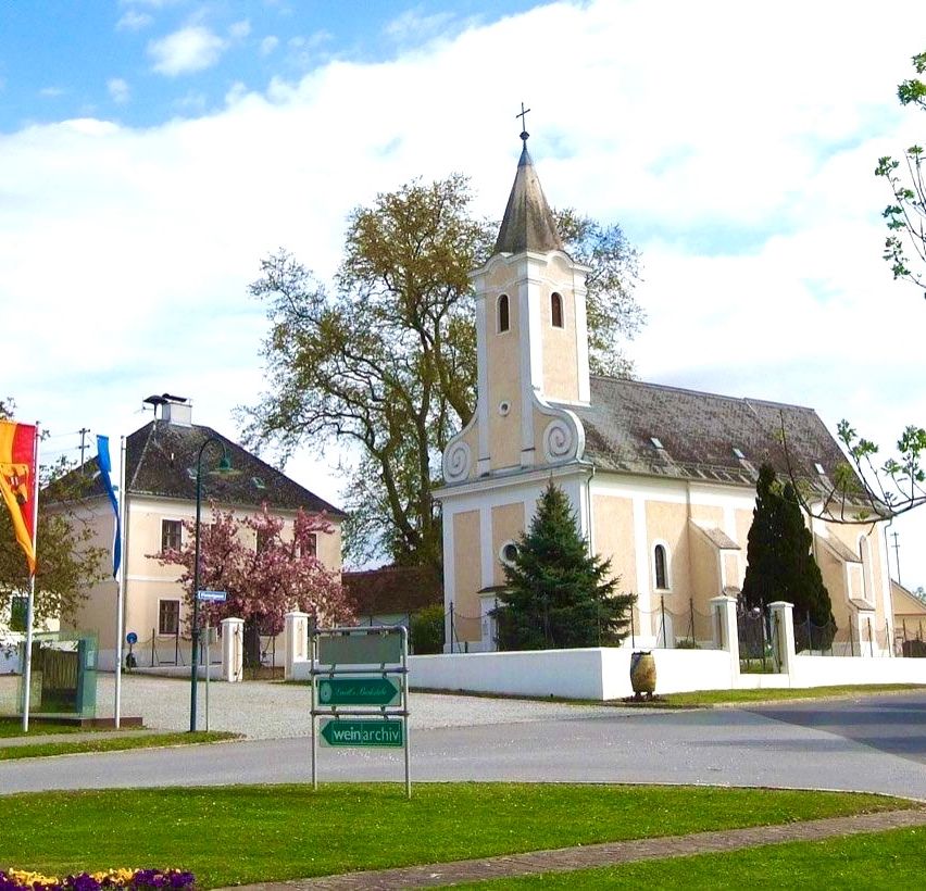 Eine Kirche mit Turm steht neben einem Gebäude mit einer roten Fahne. Straßenschilder zeigen die Richtung nach Weinarchiv. Im Vordergrund befinden sich Bäume und Blumen.