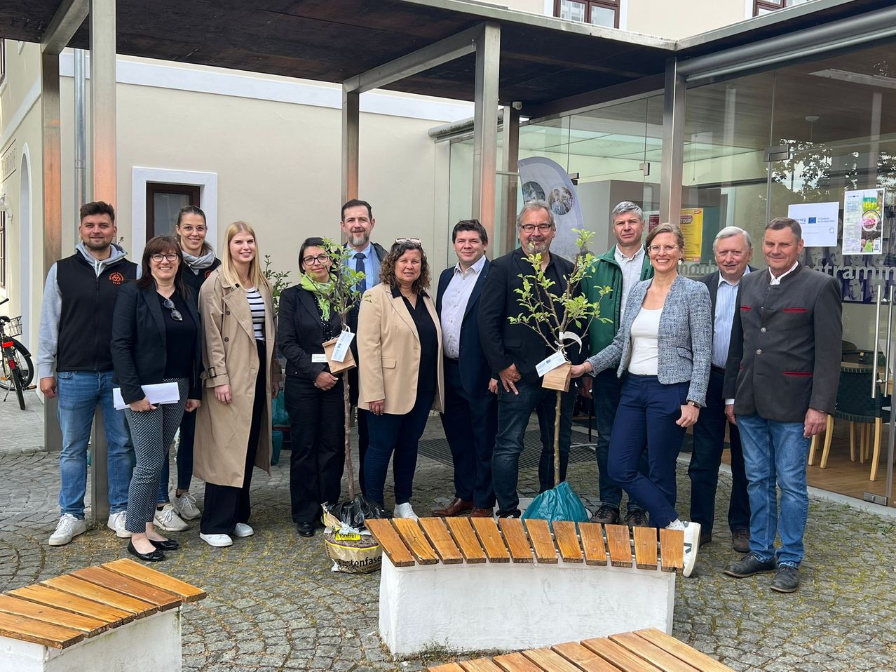 Eine Gruppe von Personen, einige mit Pflanzen in der Hand, steht vor einem Gebäude mit Vordach und Glaswand. Sie tragen formelle Kleidung und lächeln für ein Foto.