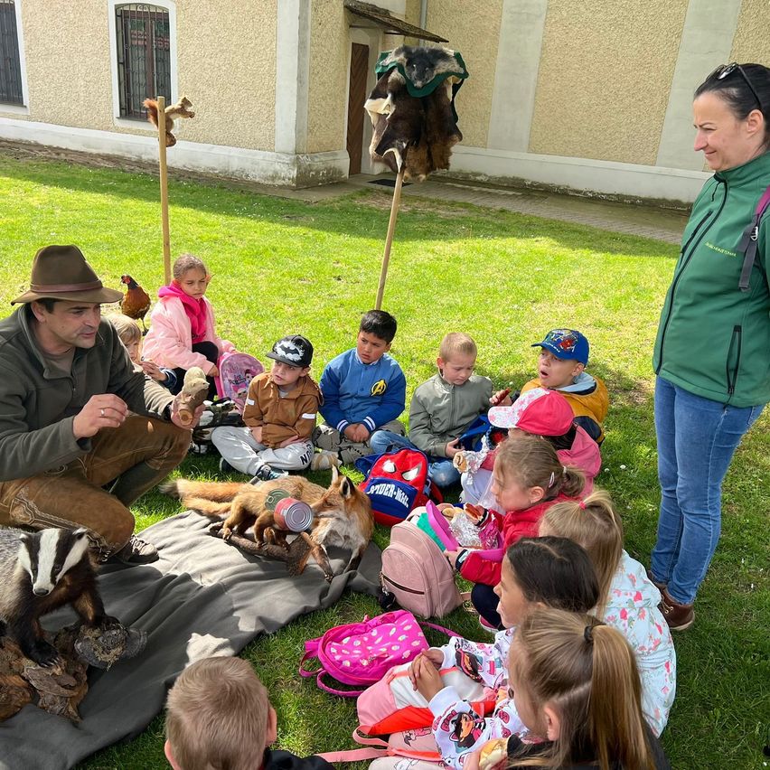 Eine Gruppe von Kindern sitzt auf dem Gras und beobachtet einen Mann, der einen Fuchs hält. Eine Frau steht in der Nähe und lächelt.
