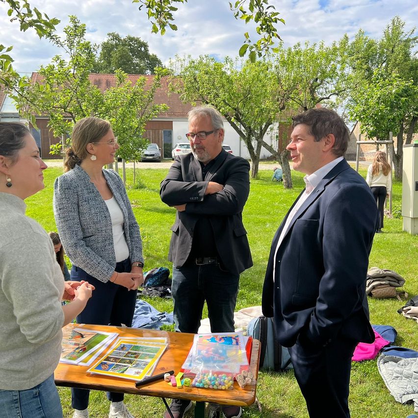 Vier Personen stehen um einen Tisch in einem Garten. Zwei Männer tragen Anzüge und zwei Frauen Blazer. Auf dem Tisch liegen Papiere, Süßigkeiten und andere Gegenstände. Hinter ihnen stehen Bäume, ein Haus und ein Auto.