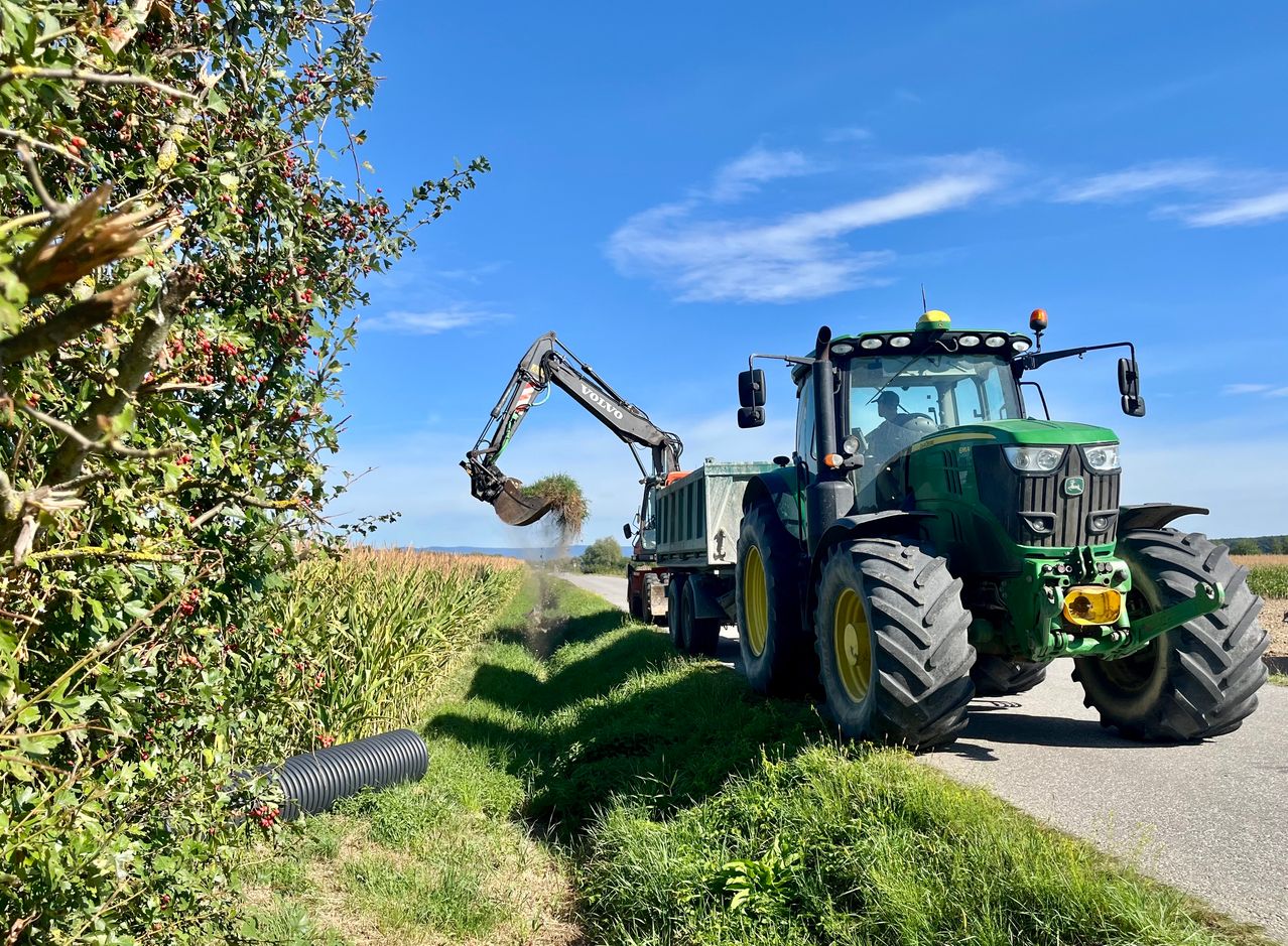 Ein grüner Traktor mit einem Volvo-Anbaugerät holt Pflanzen von der Straßenseite an einem sonnigen Tag ab.