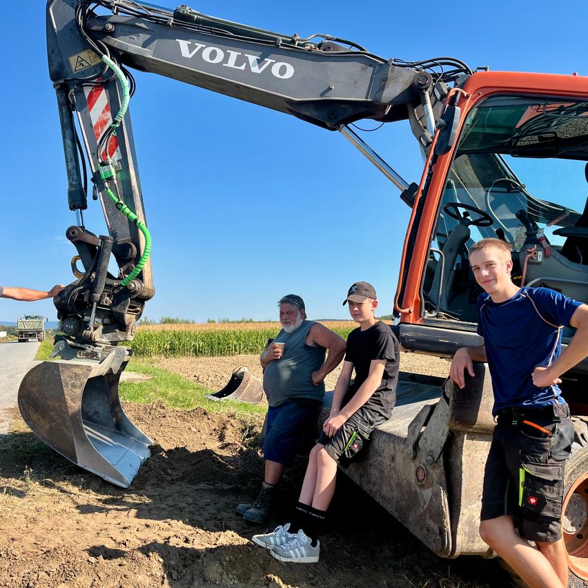Drei Männer stehen neben einem Volvo-Bagger auf einem Feld. Ein Mann hält einen Becher. Die anderen beiden tragen Mützen und Turnschuhe.