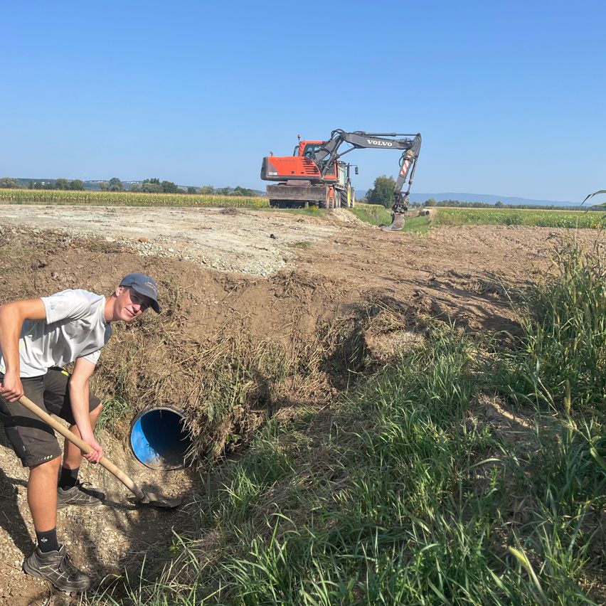 Ein Mann gräbt ein Loch mit einer Schaufel in einem Erdfeld, während im Hintergrund ein Volvo-Bagger steht.