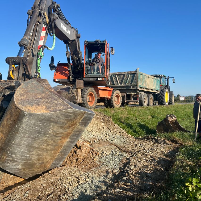 Ein Volvo-Bagger gräbt mit einem großen Schaufel in die Erde, neben einem grünen Kipplaster. Der Bediener sitzt im Bagger, und eine Person steht rechts.