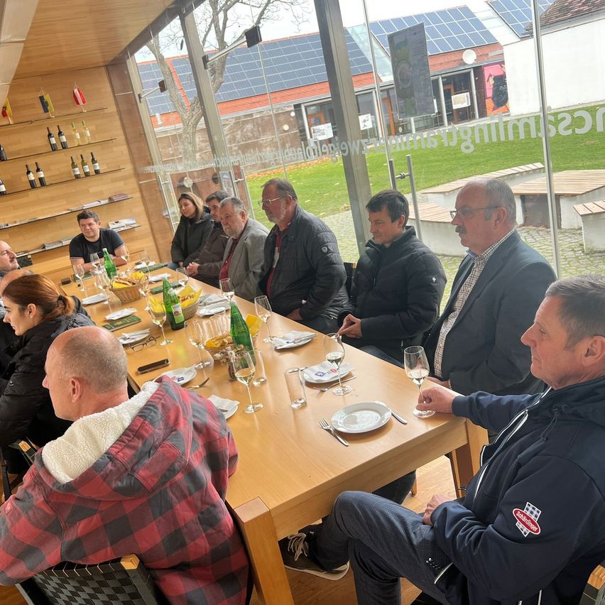 Eine Gruppe von Menschen sitzt um einen Tisch in einem Raum mit Glaswänden, die auf eine Wiese mit Solarpanelen blicken. Flaschen mit Wein sind an einer Wand aufgestellt, und ein Tisch mit Tellern, Gläsern und Weinflaschen ist gedeckt.