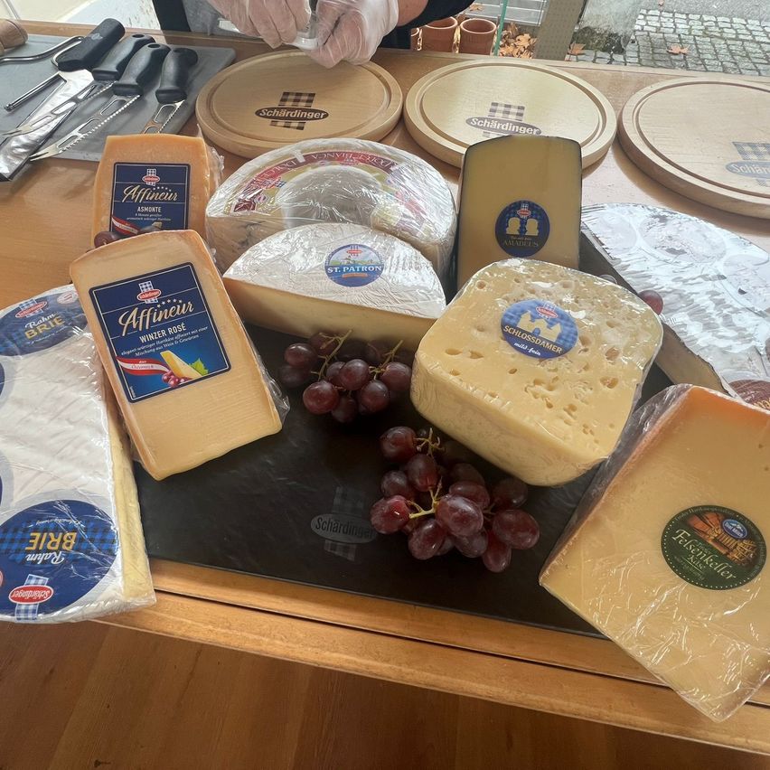 Eine Auswahl verschiedener Käsesorten auf einem Holztisch mit Holzschneidebrettern, Messern und Trauben. Bemerkenswerte Käsesorten sind Affineur, St. Patron und andere.