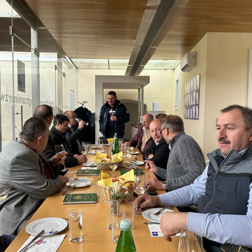 Eine Gruppe von Menschen sitzt um einen langen Tisch mit Weingläsern und Tellern und genießt eine Mahlzeit. Ein Mann steht in der Nähe einer Tür, und andere schauen zu ihm. Der Tisch ist mit Brot, Käse und Weinflaschen gedeckt.